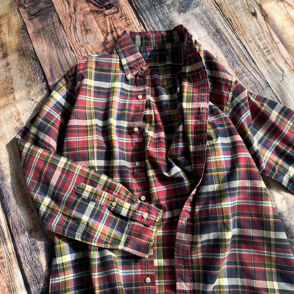 木目の床に平置きされた、ラルフローレンの赤・黄・青のマルチカラー・チェックシャツ。洗いざらしの風合いが残るアメカジらしい一着。